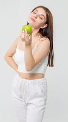 Cute young brown-haired girl tries a green apple on a white background/The model is wearing a white top and knitted pants