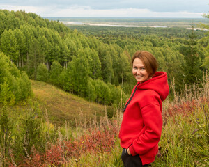 Fototapeta na wymiar a smiling woman with red hair and a red hoodie on a wooded hill against the background of a forest and a winding river in the distance.