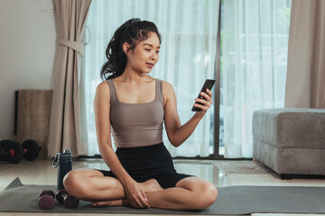 Asian woman exercise at home. Healthy female in sportswear sitting and using smartphone in living room, Health care and wellness concept.