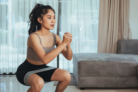 Asian woman exercise at home. Healthy female in sportswear workout doing squat with rubber band in living room, Health care and wellness concept.