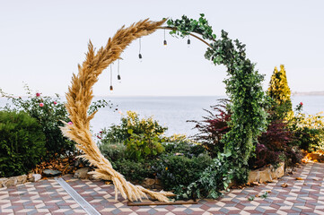 A beautiful round wedding arch, decorated with reeds, green leaves of plants, with electric hanging...
