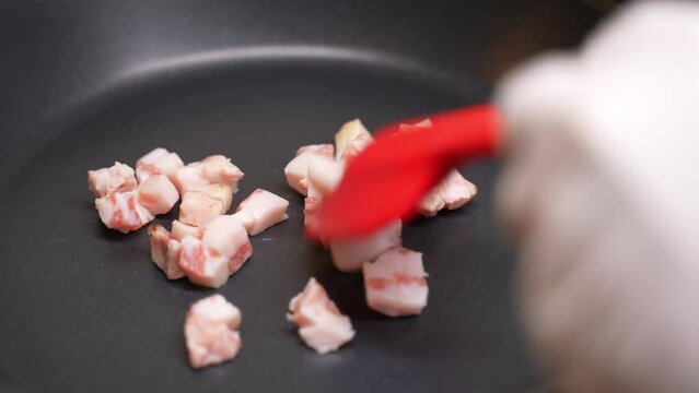 Closeup slow motion of cook or chef frying pieces of fresh bacon in cubes into a frying pan.