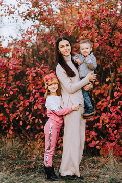 Photography, a portrait of a beautiful brunette girl with a small child, a boy, a son, and a smiling girl, a daughter, in nature in autumn, against the backdrop of a tree, bushes with red leaves.