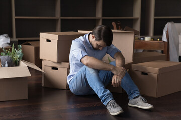 Upset frustrated man sit on floor near heap of stacked cardboard boxes with stuff feels depressed,...
