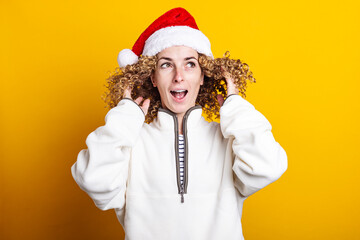 Cheerful curly young woman in santa claus hat on a yellow background.