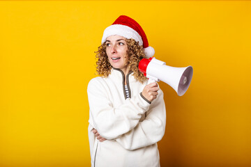 Young woman in santa claus hat with a megaphone on a yellow background.