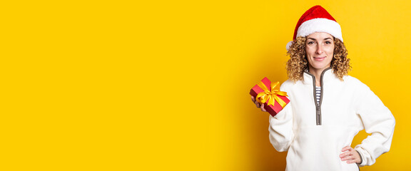Smiling young woman in santa claus hat holding a gift on a yellow background. Banner.
