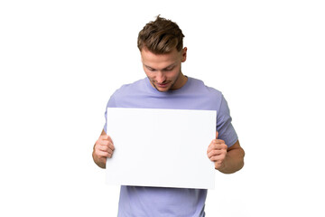 Young blonde caucasian man over isolated background holding an empty placard and looking it