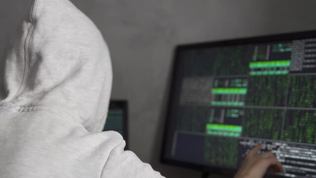 Hooded IT programmer working on desktop computer in data center technical system control room. Young hacker working with computer, close up.