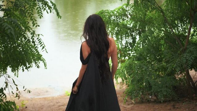 Fantasy gothic woman queen in long elegant silk black dress walks along river bank. Portrait mystic girl princess rear view looks back. Green trees forest summer nature, lake water. Lady vampire witch