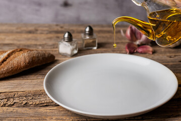 Extra virgin olive oil pouring from an oil can into a round white plate, on a rustic wooden table, with bread, salt and pepper.
