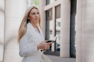Thoughtful young adult businesswoman holding diary dressed in white suit looks up, makes decision...