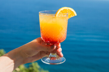 Slightly alcoholic drink with a slice of orange in the hands woman against the backdrop of the blue sea.