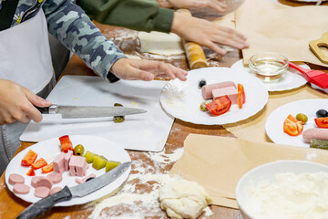 Cropped children's hands with knife cut vegetables on white plastic cutting board. Nearby are products needed to make pizza. Culinary training.