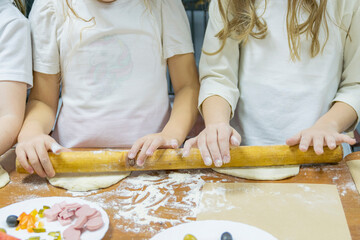 Two girls use one rolling pin to roll out two pieces of dough at same time on wooden table. Cooking training - first pizza preparation.
