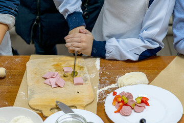 Cropped children's hands with knife cut vegetables and sausage on wooden cutting board. Nearby are products needed to make pizza. Culinary training.