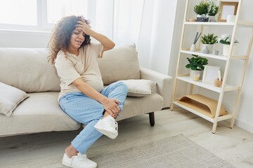 Obraz premium Pregnant woman sitting on sofa at home and putting on pregnancy-friendly shoes with problem due to belly, headache, difficulties of pregnancy and motherhood