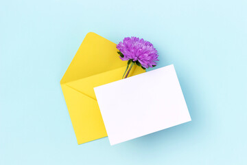 White blank card, yellow envelope and purple flower on blue background. Minimal style. Top view Flat lay Mockup