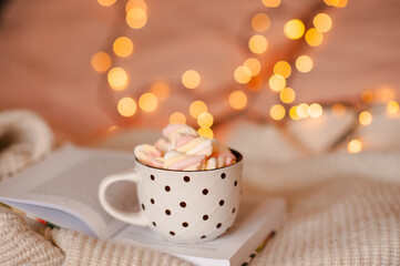Obraz na płótnie Canvas Cup of hot chocolate with marshmallow stay on open paper book in bed over Christmas lights close up. Winter greeting holiday season.