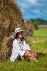 Naklejka na ściany i meble Portrait of an young gorgeous woman without makeup wearing a white shirt, black shorts, and a hat. Beautiful country girl resting on a summer field. Haystack in the background.