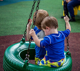 two cute young boys, brothers playing on tire swing in park with playground turf in background