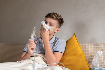 Sick teen boy with an inhaler. Unhealthy child doing inhalation at home, she use nebulizer and inhaler for the treatment, sitting on the couch at home. Asthma inhaler, nebulizer steam, flu or cold