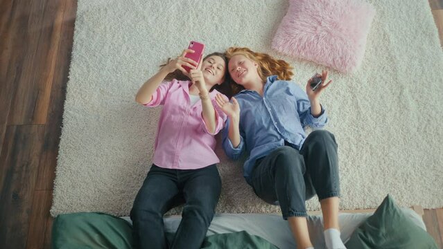 Two Teenage Girls Having Fun While Lying on the Floor at Home. Uses a Smartphone, Sends Text Messages, Making a Selfie, Chatting. Adolescence Concept, Emotional Health. Body Positive. Mental Health.