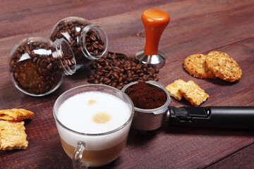 Cup of cappuccino, crackers, holder with ground coffee, tamper and coffee beans on wooden table.