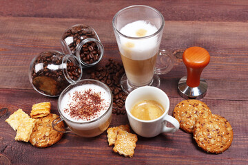Cups of espresso, latte and cappuccino, crackers, holder with ground coffee, tamper and cans of coffee beans on wooden table.