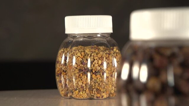 Close up refocusing zoom out and in of two plastic jars with perga and pollen on table. Jars with white caps. Background is darked and out of focus