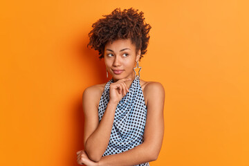 Beautiful curly haired woman keeps hand under chin touches jawline thinks about something ponders...