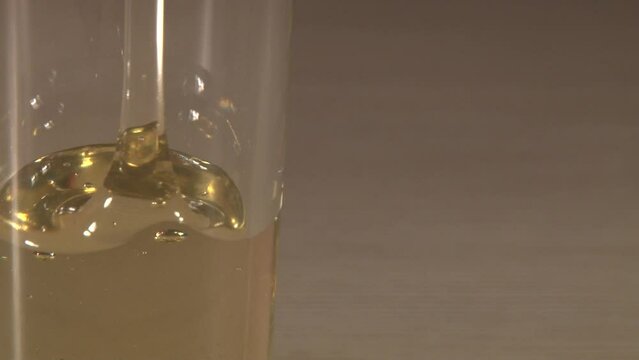 Close up of transparent glass standing on the table with pouring golden honey shooted from side trough the glass. Artifical lighting indoor