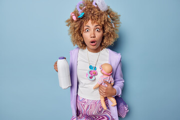 Young shocked woman anticipates for baby holds bottle of milk and doll wears casual clothes string with nipples sees something with astonishment isolated over blue wall. Expectant mother indoors