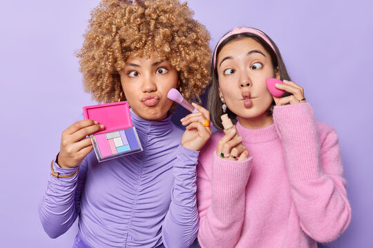 Two funny women do makeup apply eyeshadow and foundation make funny grimace fish lips prepare for party want to have fabulous look dressed casually isolated over purple background. Beauty procedures