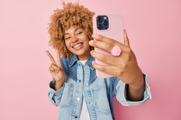 Good looking cheerful woman makes peace gesture smiles broadly clicks selfie via smartphone wears denim jacket enjoys spare time isolated over pink background records video for chat makes photo - Powered by Adobe