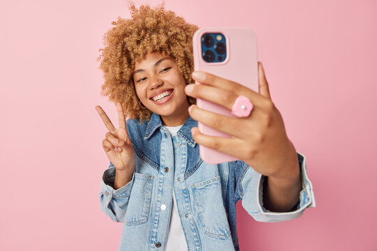 Good looking cheerful woman makes peace gesture smiles broadly clicks selfie via smartphone wears denim jacket enjoys spare time isolated over pink background records video for chat makes photo