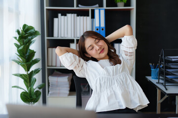 Young Asian businesswoman stretches her arms and closes her eyes to relax her tired muscles from working at her desk all day at the office.