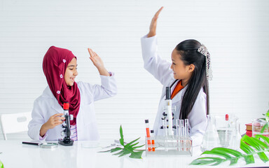 Two young teenage diverse student girls wearing white gown, smiling with fun, studying chemistry,...