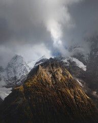 Dramatic view of forested high rocky slopes and snowy mountains in gray cloudy sky in changeable weather, mountain landscape without people