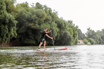 Man on stand up paddle board (SUP) paddling along the calm summer Danube river against a background of green trees at the riverside