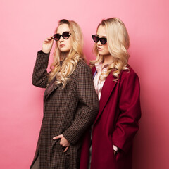 Fashionable two blond women in coat with sunglasses posing on pink background. Fashion autumn winter photo.