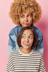 Vertical shot of beautiful thoughtful diverse young women friends purse lips and look pensively aside wear casual clothes focused aside being deep in thoughts isolated over pink studio background