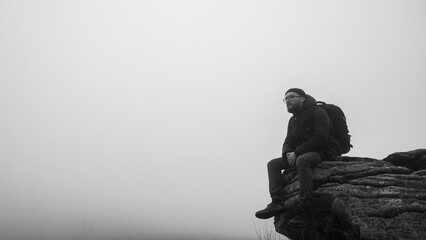 a young man in glasses and a beard, a traveler, sits on the edge of a mountain, fog, mist, rest in the mountains. Free space for writing	
