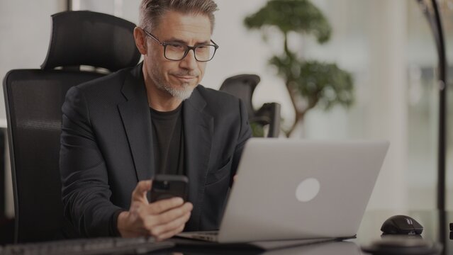 Business Portrait - Businessman Working With Phone And Laptop Computer In Corporate Office. Happy Middle Aged, Mid Adult, Mature Age Man Smiling.