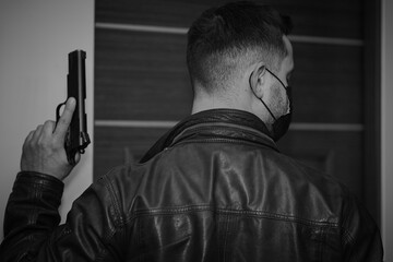 A man in a leather jacket and a mask on his face with a gun in his hand, view from the back.