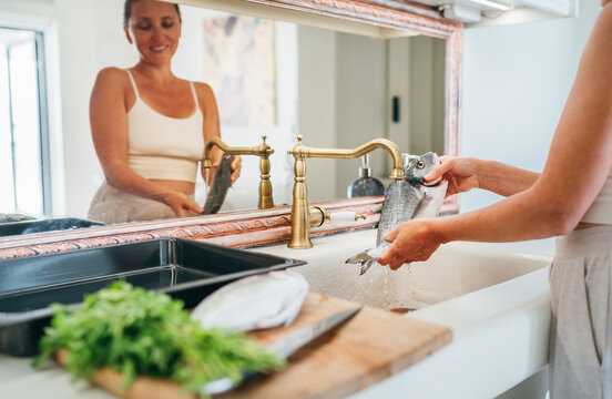 Smiling young adult woman washing fish under the kitchen sink water tap. Healthy seafood preparation and house work concept image.