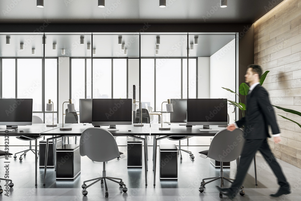 Poster businessman walking by workspace tables with modern computers in spacious coworking office with wood