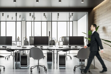 Businessman walking by workspace tables with modern computers in spacious coworking office with wooden wall and panoramic window