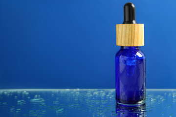 Bottle of face serum on wet surface against blue background, closeup. Space for text