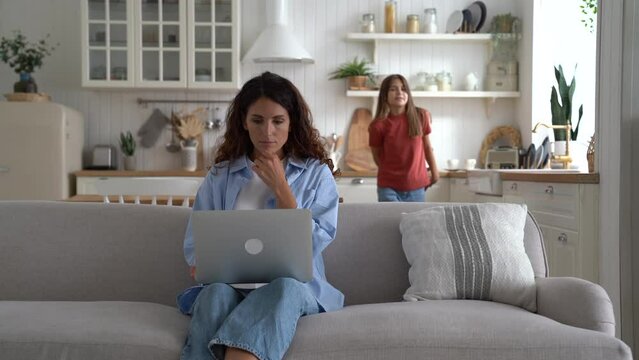 Pleased girl child makes surprise gives gift to mother who works hard with computer on couch at home. Happy family concept. Contented cute kid daughter making present on mothers day for young mommy.
